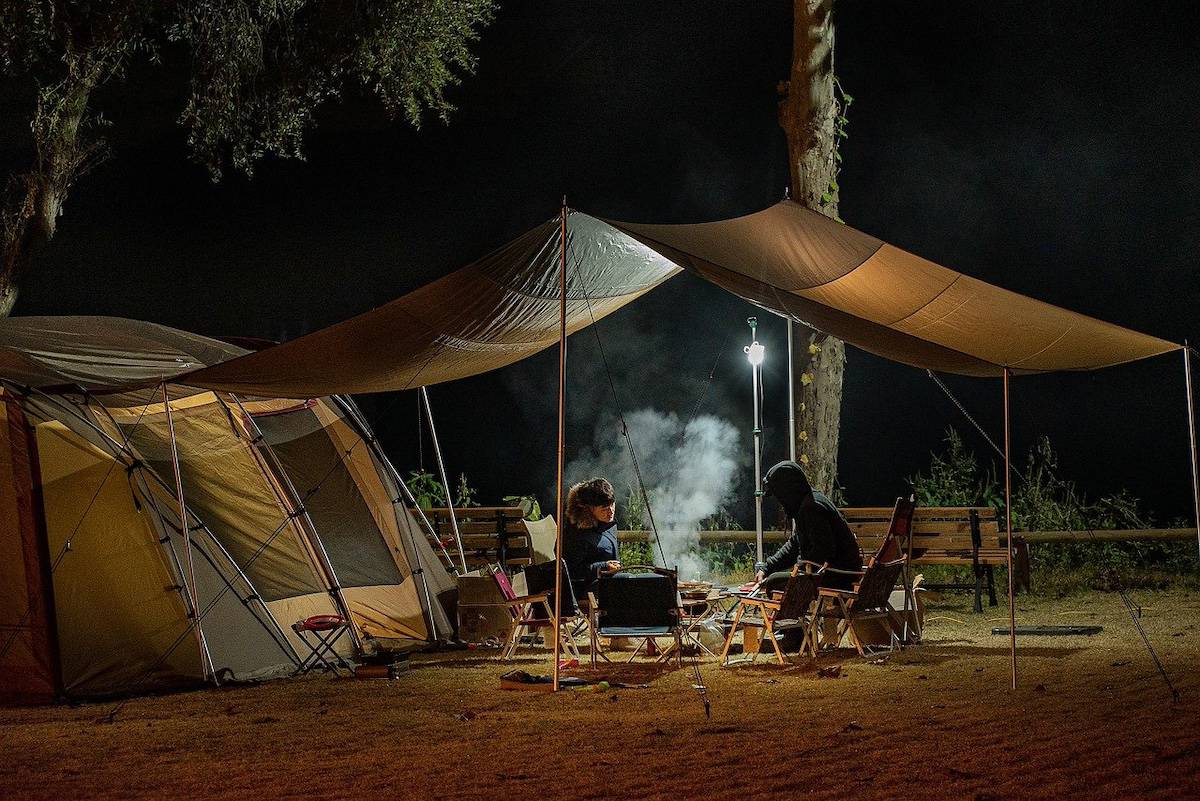Two warmly dressed people sitting under the flap outside their tent, cooking food on a braai at night