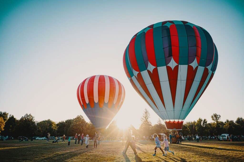 Two Hot air balloons landing in a grassy field with a lot of sunshine and people around.