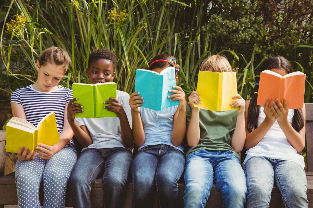 5 Children sitting on a bench in the sunshine reading, against a background of of tall grass