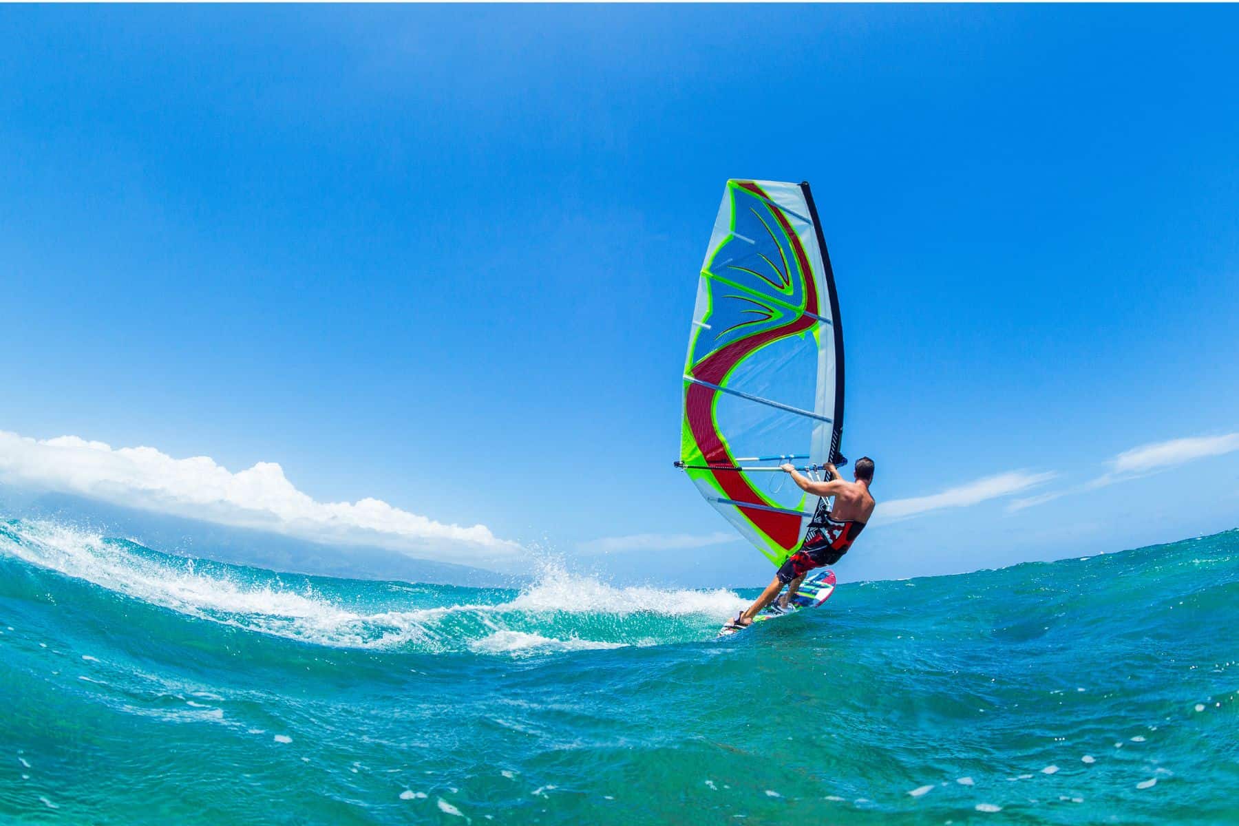 A man on a colourful windsurfer in the calm blue water, against an sunny sky