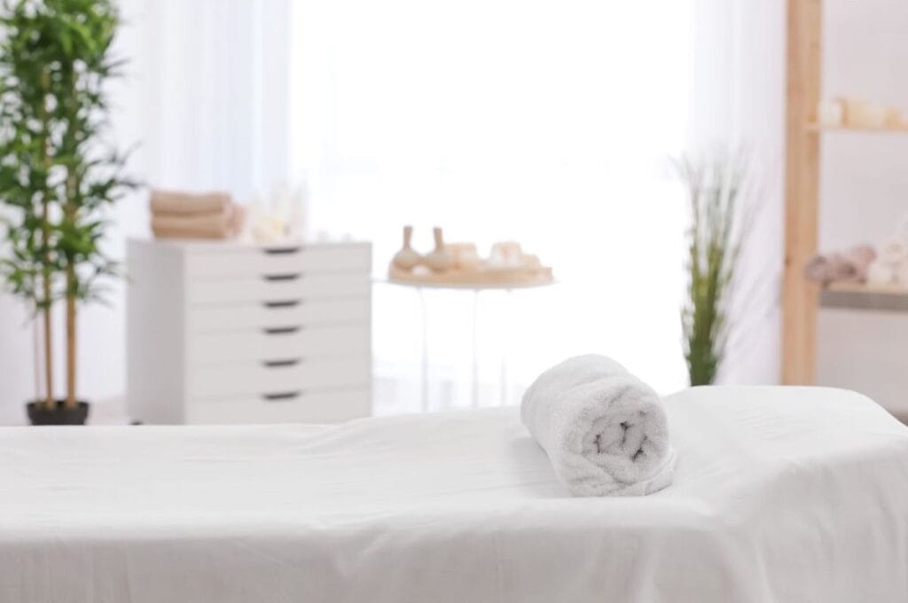 A bright white room with a spa table covered in white. A rolled towel sits on top, and a tall green plant stands in the corner.