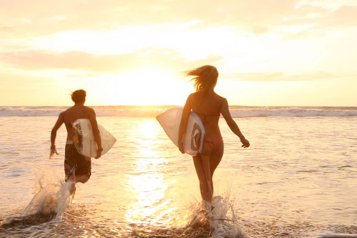 Two people running into the waves with their surfboards as the sun goes down