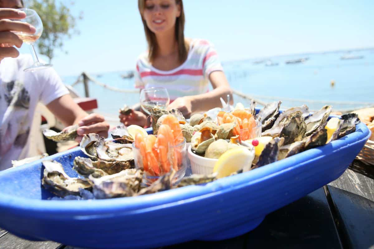 A couple dining on a tray of oysters and shrip with the sea in the background under a bright blue sky. DP